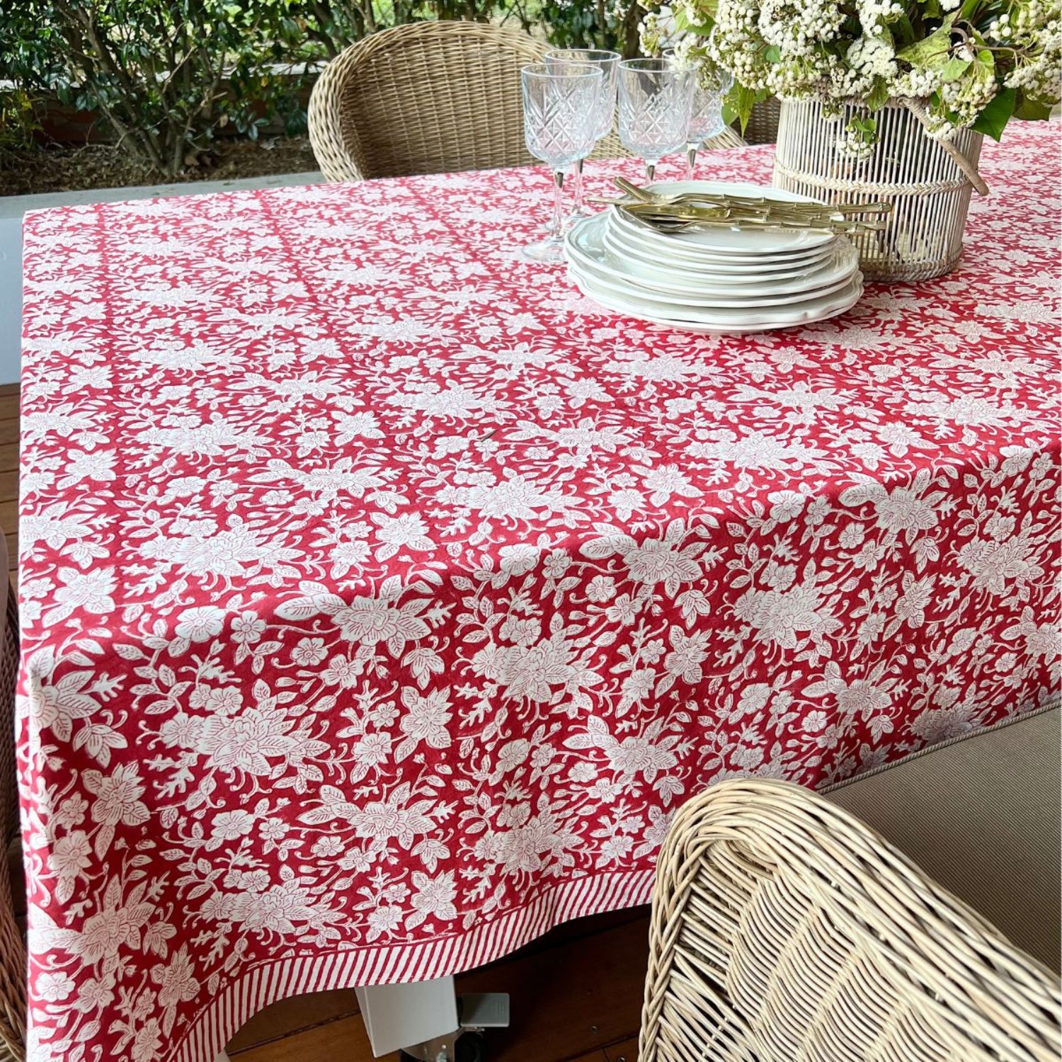 A red and white hand block printed floral patterned square tablecloth on a table with plates and cutlery set up for a meal.