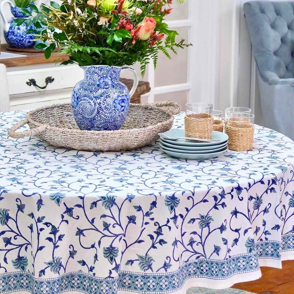 A round tablecloth with a blue floral hand block printed pattern displayed on a table accompanied by a blue and white ceramic vase and glass candle holders.