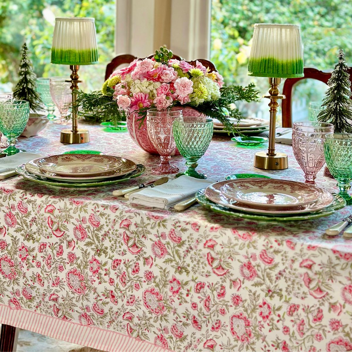 A set dining table featuring a pink and green floral patterned tablecloth, with matching napkins, glasses, and a floral centerpiece.