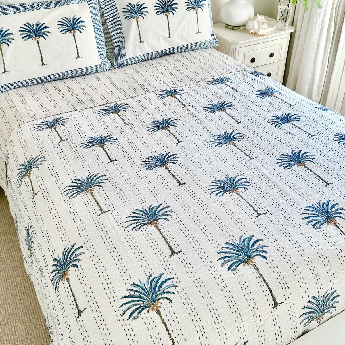 A hand block printed bedcover with a blue and white palm patterned Kantha quilt, with pillows matching the bedspread, in a room with light decor.