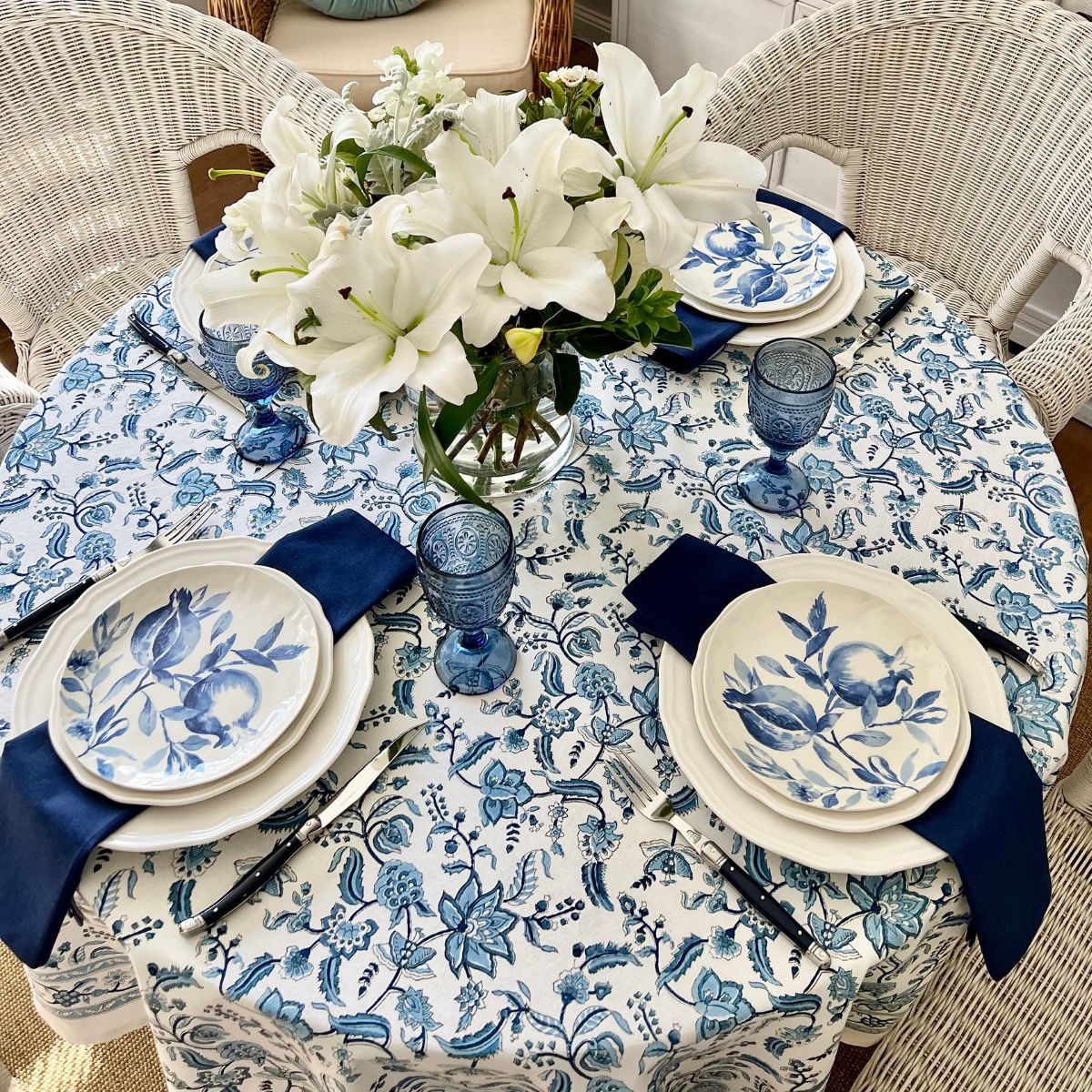 A 152 cm Chintz monochrome blue hand block printed cotton round tablecloth on a 4 seater circular dining table, styled for a relaxed casual meal with white and blue dishes, a floral centrepiece, and navy blue napkins.