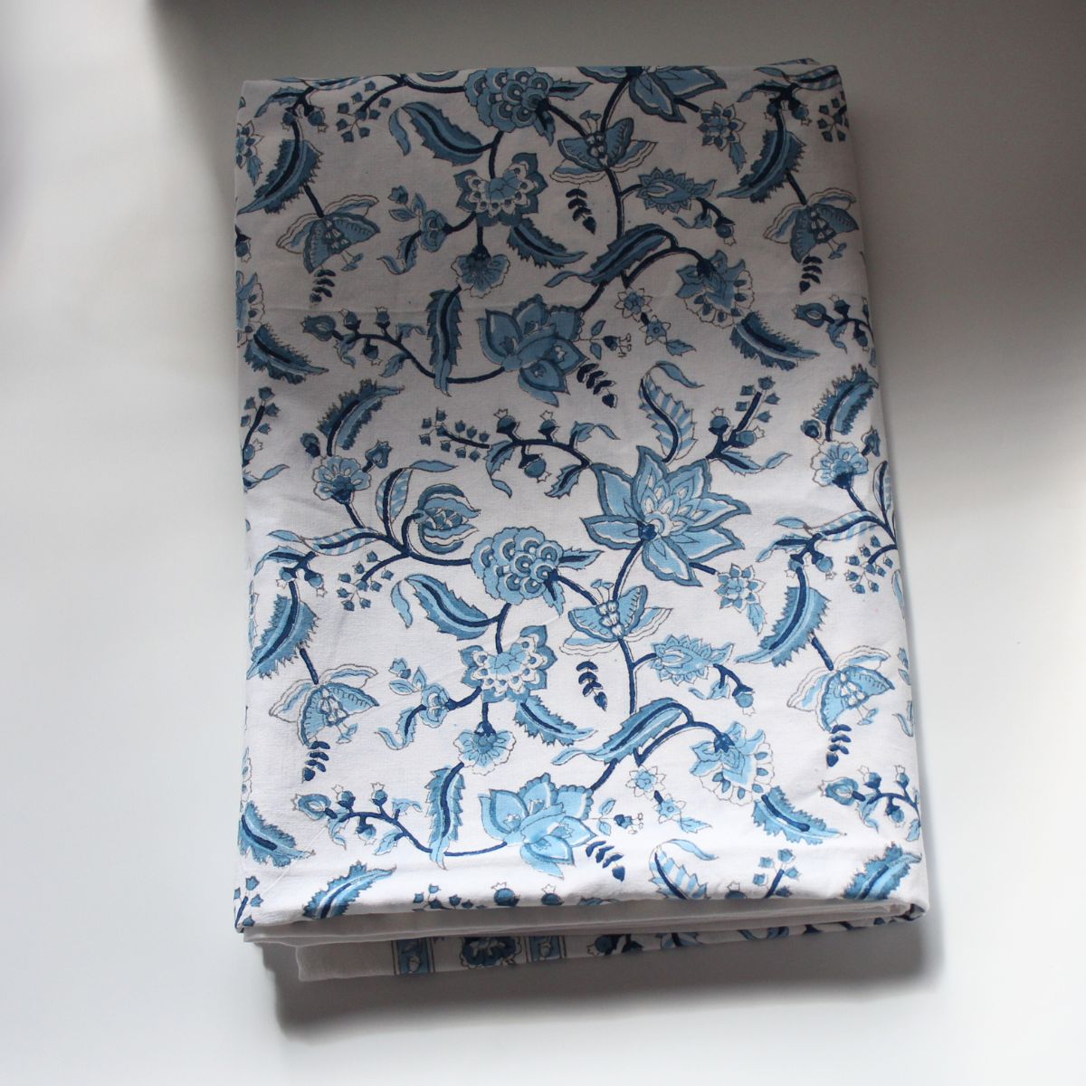 A Chintz monochrome blue hand block printed cotton square tablecloth folded flat, showing the intricate floral pattern and rich decorative border on premium cotton fabric.