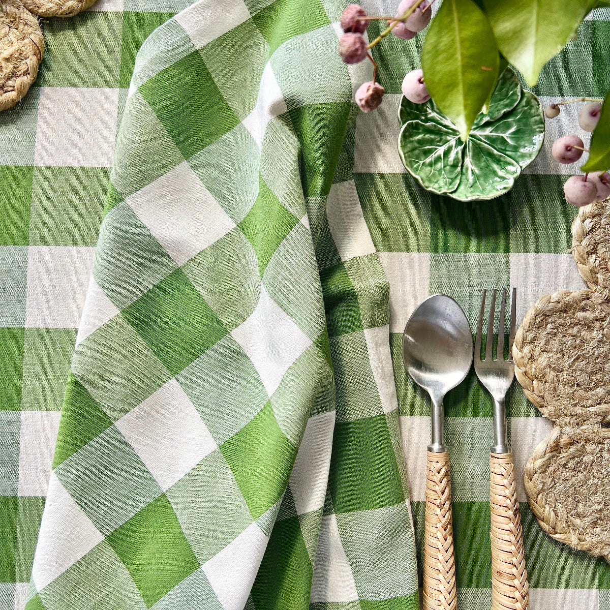 A green gingham handwoven circular tablecloth on a garden dining table set for a relaxed get-together, with decorative plates, bamboo cutlery, green gingham napkins, and a fresh floral centrepiece bringing a garden-party feel.