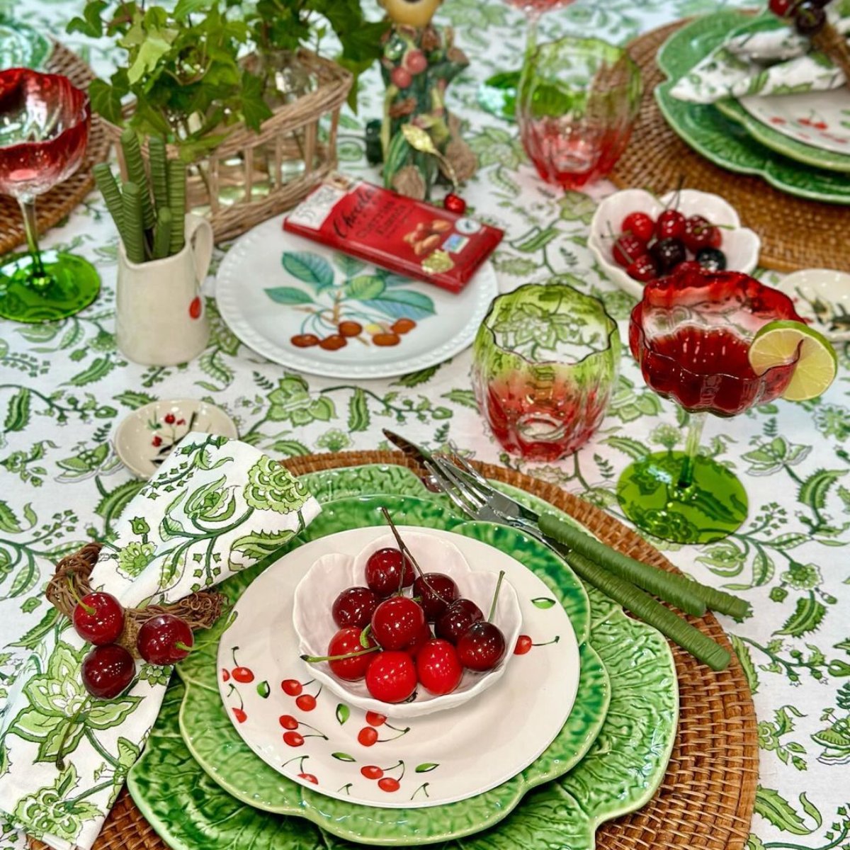 A green chintz hand block printed cotton napkin as part of a cozy everyday dining setting, the rich green chintz floral motif adding a fresh botanical touch to the table, styled with red cherrirs for a summer lunch tablescape.