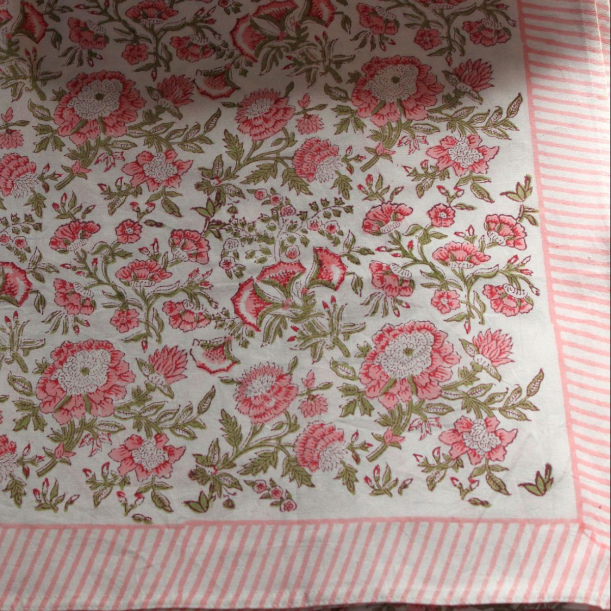 Flat-lay of a square pink and green Beatrice block print tablecloth, photographed on a neutral background to showcase the full repeat pattern and striped edge.