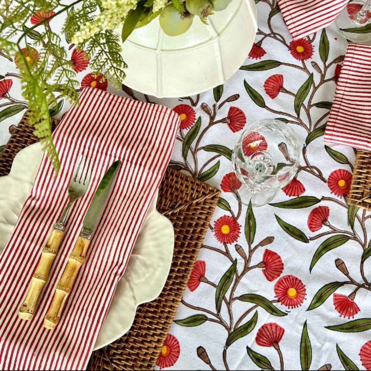 A set of red striped hand block printed table napkins with a floral pattern on the tablecloth, displayed alongside a glass of water and decorative greenery.