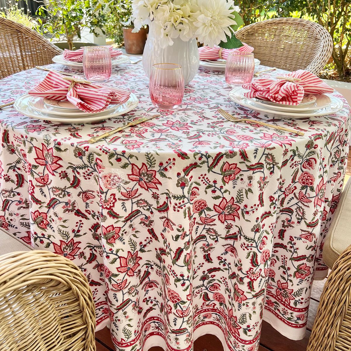A multicolored round tablecloth with a pattern of hand block printed flowers, pomegranates and green leaves, displayed on a table set with plates and glasses.