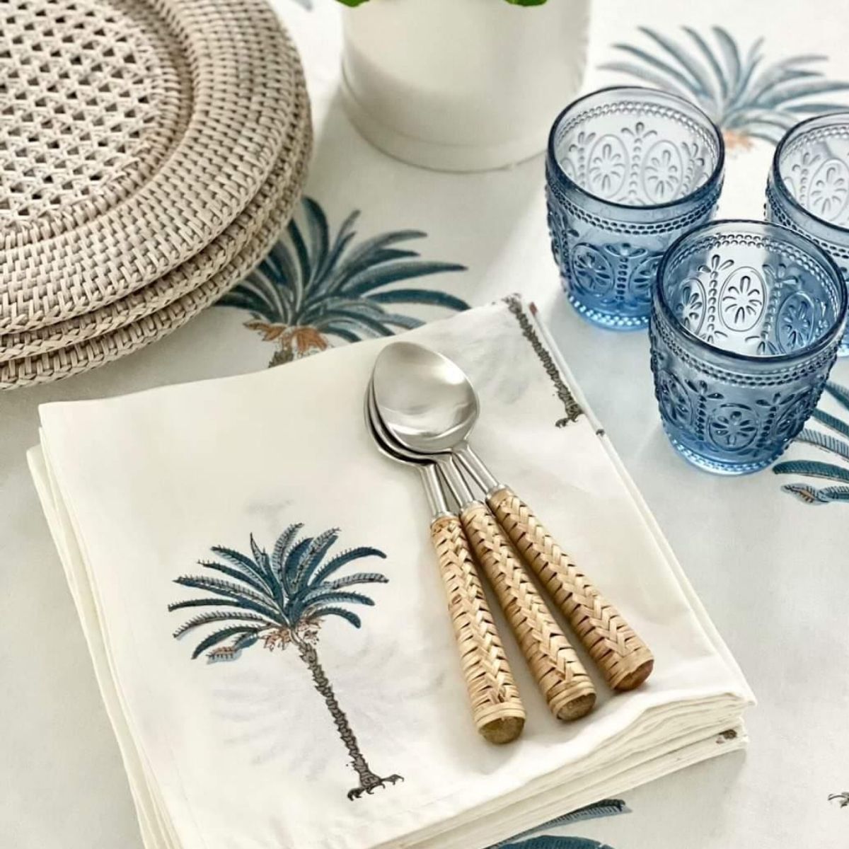 A set of white cotton napkins with a hand block printed blue palm design, displayed on a table with other tableware.