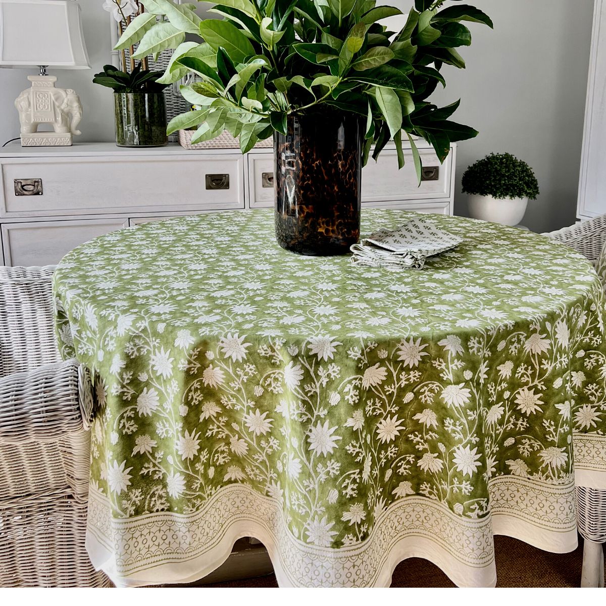 A green round tablecloth with a floral hand block printed Australian flannel pattern displayed on a table in a room with a plant and a framed picture in the background.