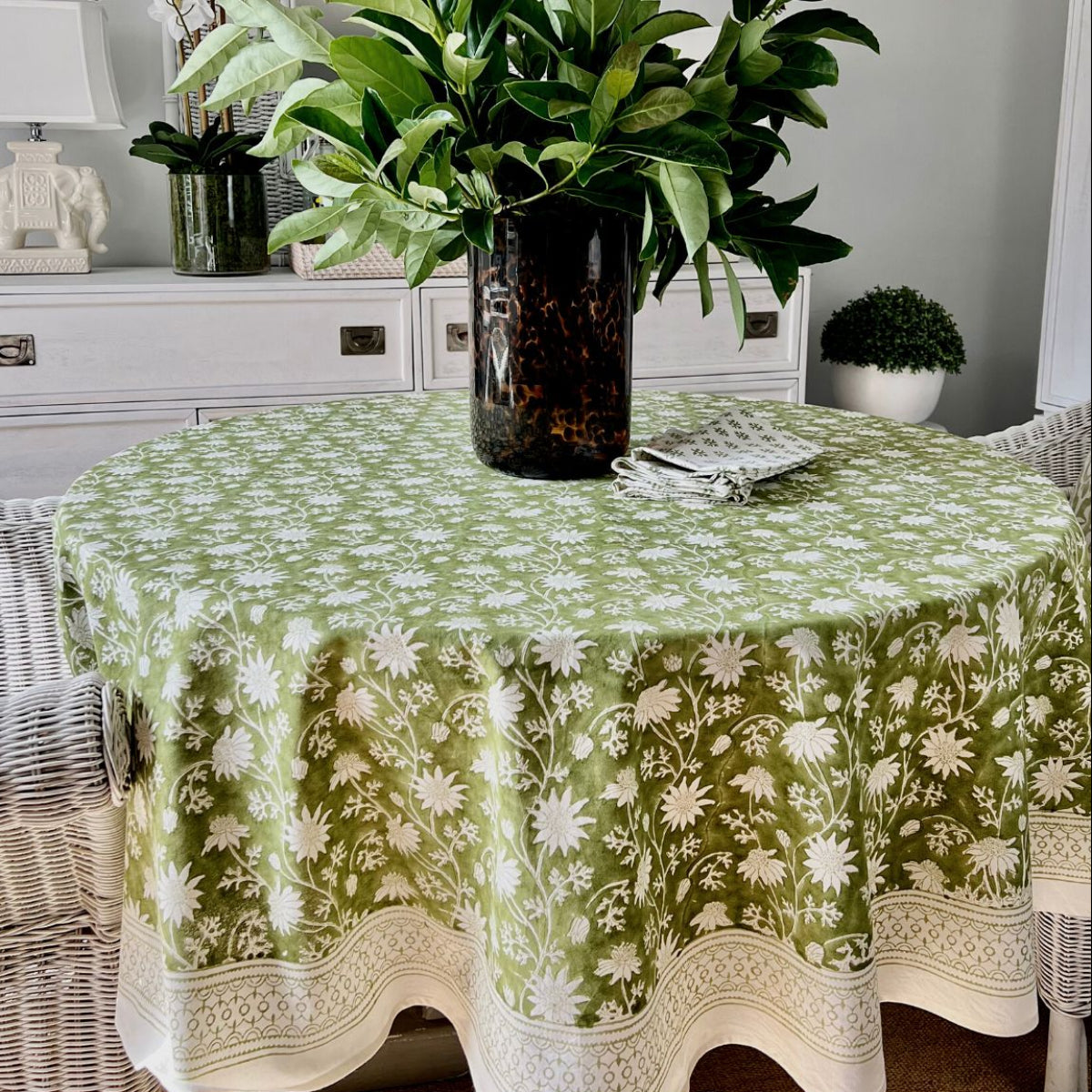 A green round tablecloth with a floral hand block printed Australian flannel pattern displayed on a table in a room with a plant and a framed picture in the background.