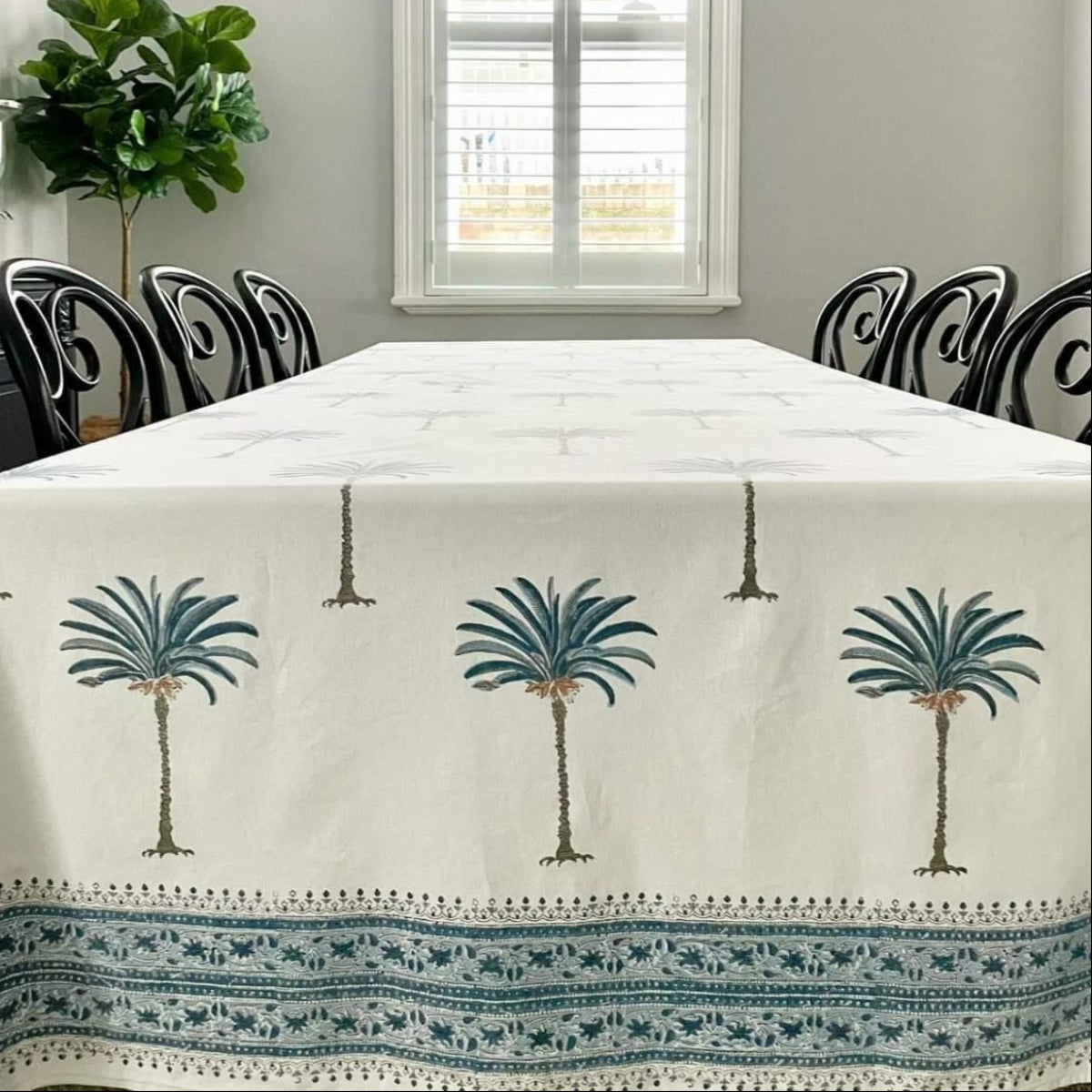 A rectangular hand block print tablecloth with a blue palm motif pattern on cotton fabric, laid out on a table with black chairs.
