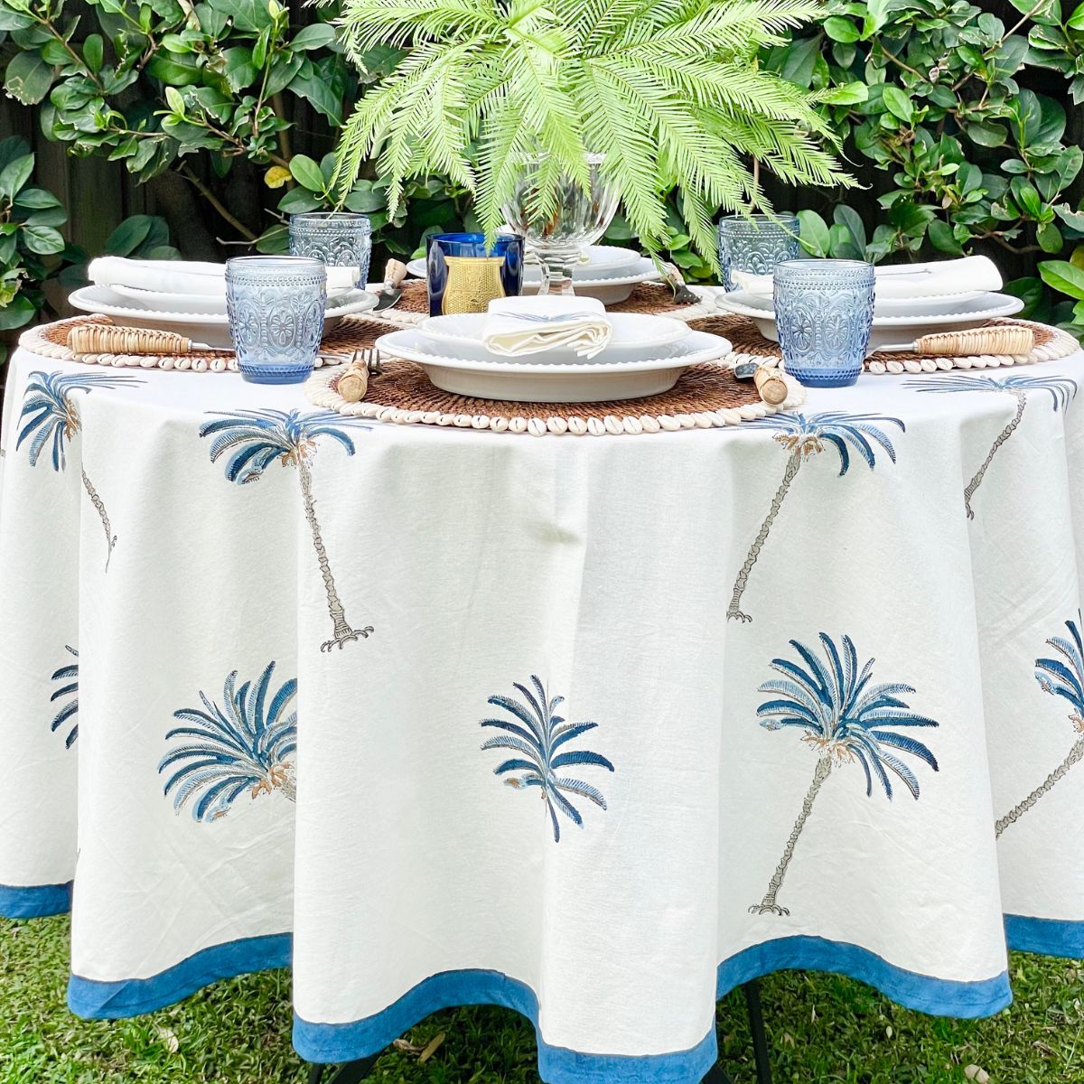 A round hand block printed tablecloth with a blue palm tree pattern displayed on a table outdoors, surrounded by plants.