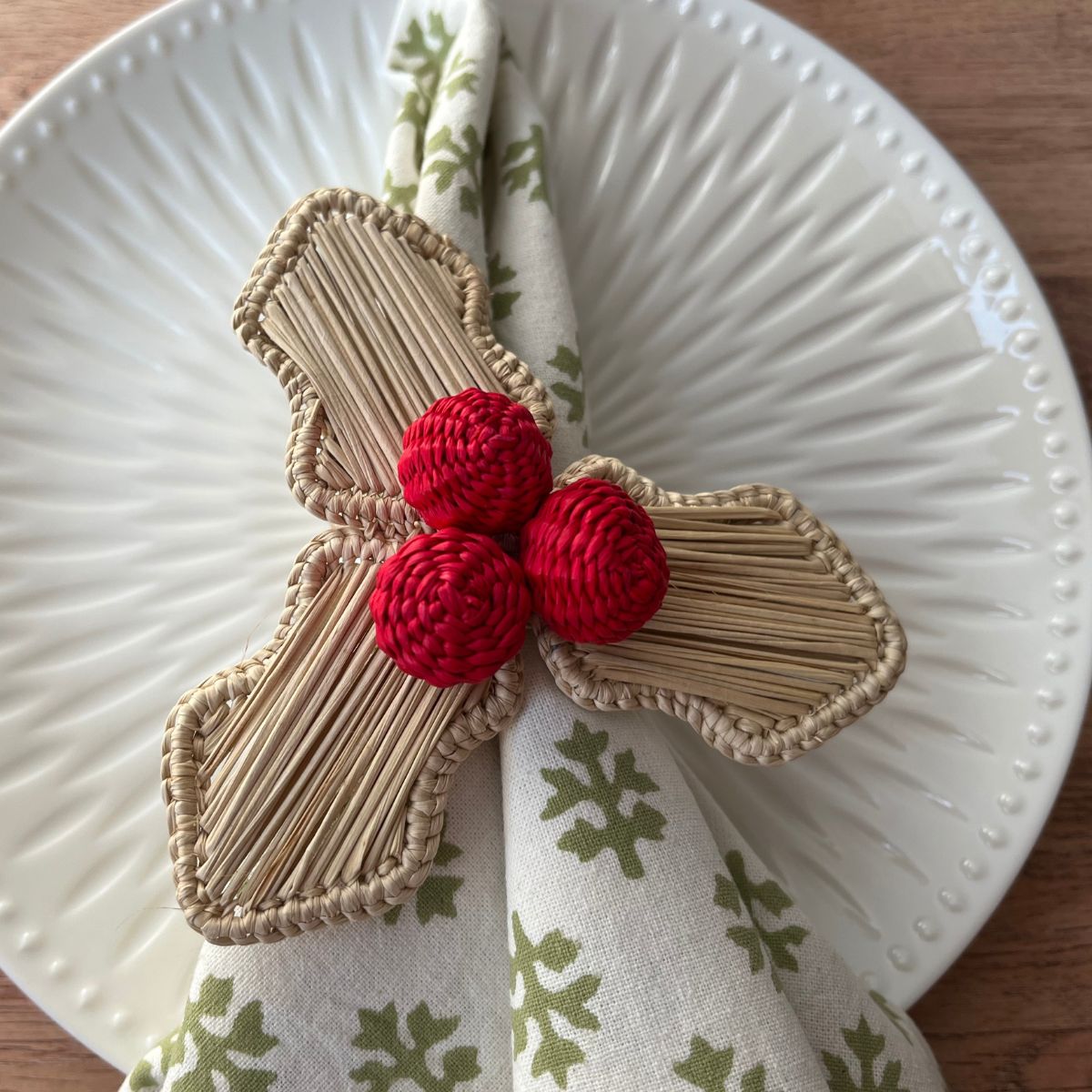 A red napkin ring with a holly berry design, consisting of red and beige accents, displayed on a white plate with a green patterned napkin.