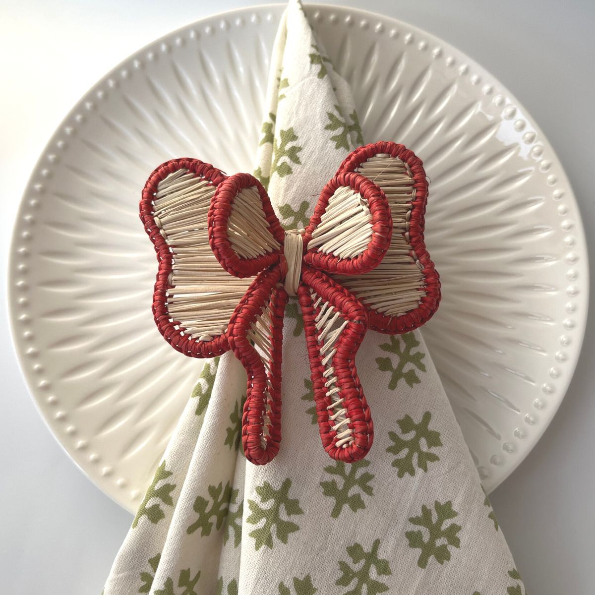 A red bow napkin ring on a table napkin with a green leaf pattern, displayed on a table setting with white plates and cutlery.