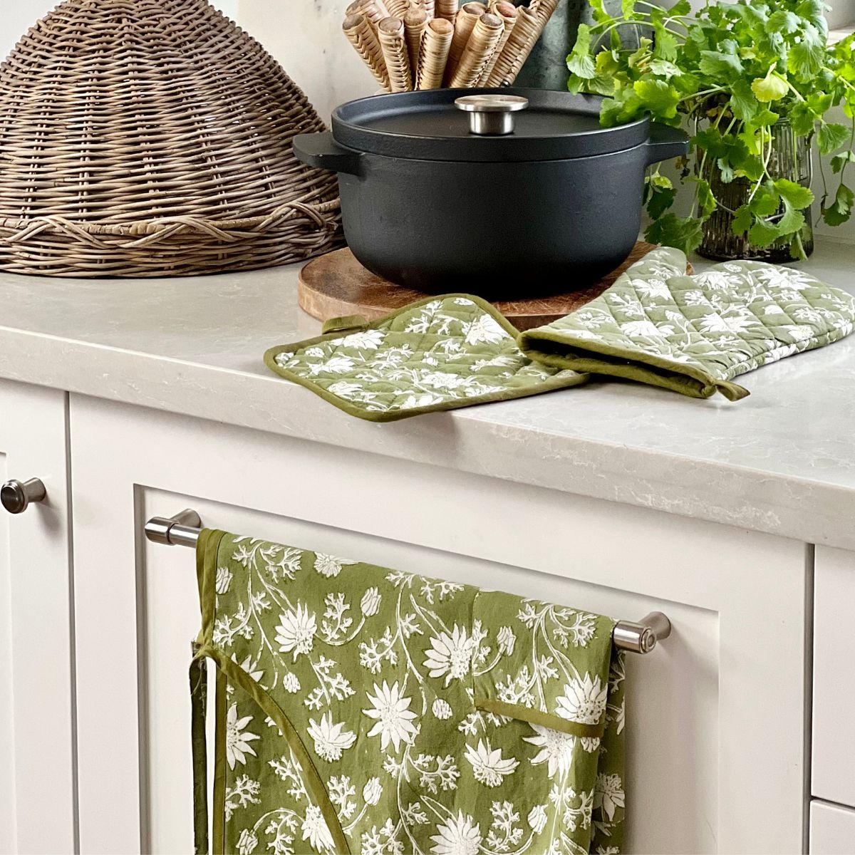 Flannel Flowers apron, oven mitt, and pot holder styled together beside cookware, creating a coordinated artisan kitchen linen collection for cooking, baking, and gifting.