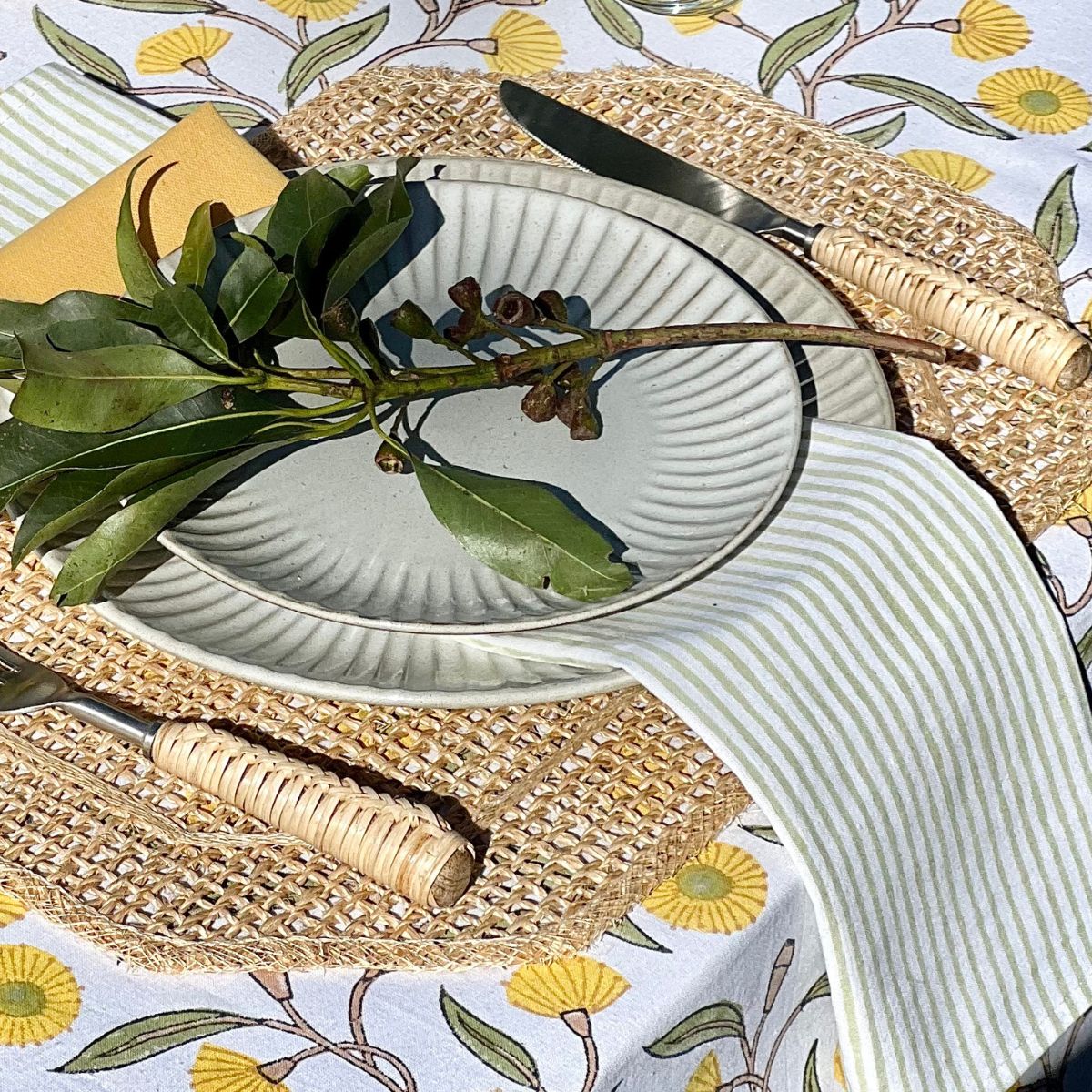 A green striped cotton napkin displayed over white plates on a yellow and green floral tablecloth, perfect for everyday meals and outdoor entertaining.