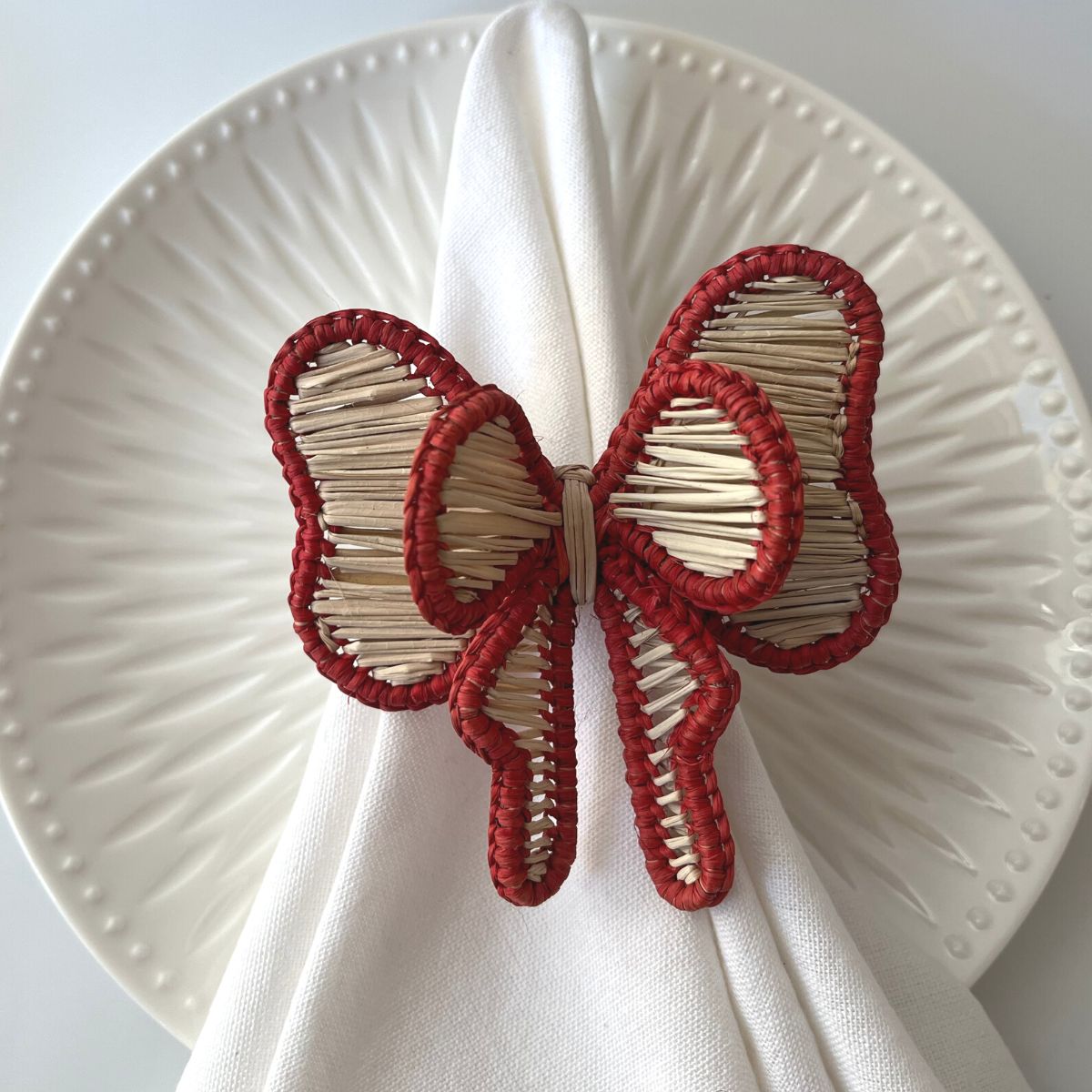 A red bow-shaped napkin ring made from natural straw, displayed on a white plate with a napkin.