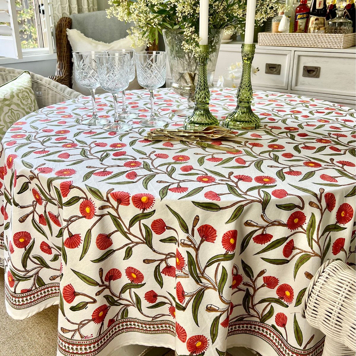 A round tablecloth with a red green flowering gum hand block printed design displayed on a table, paired with glassware and a white wicker chair in the setting.