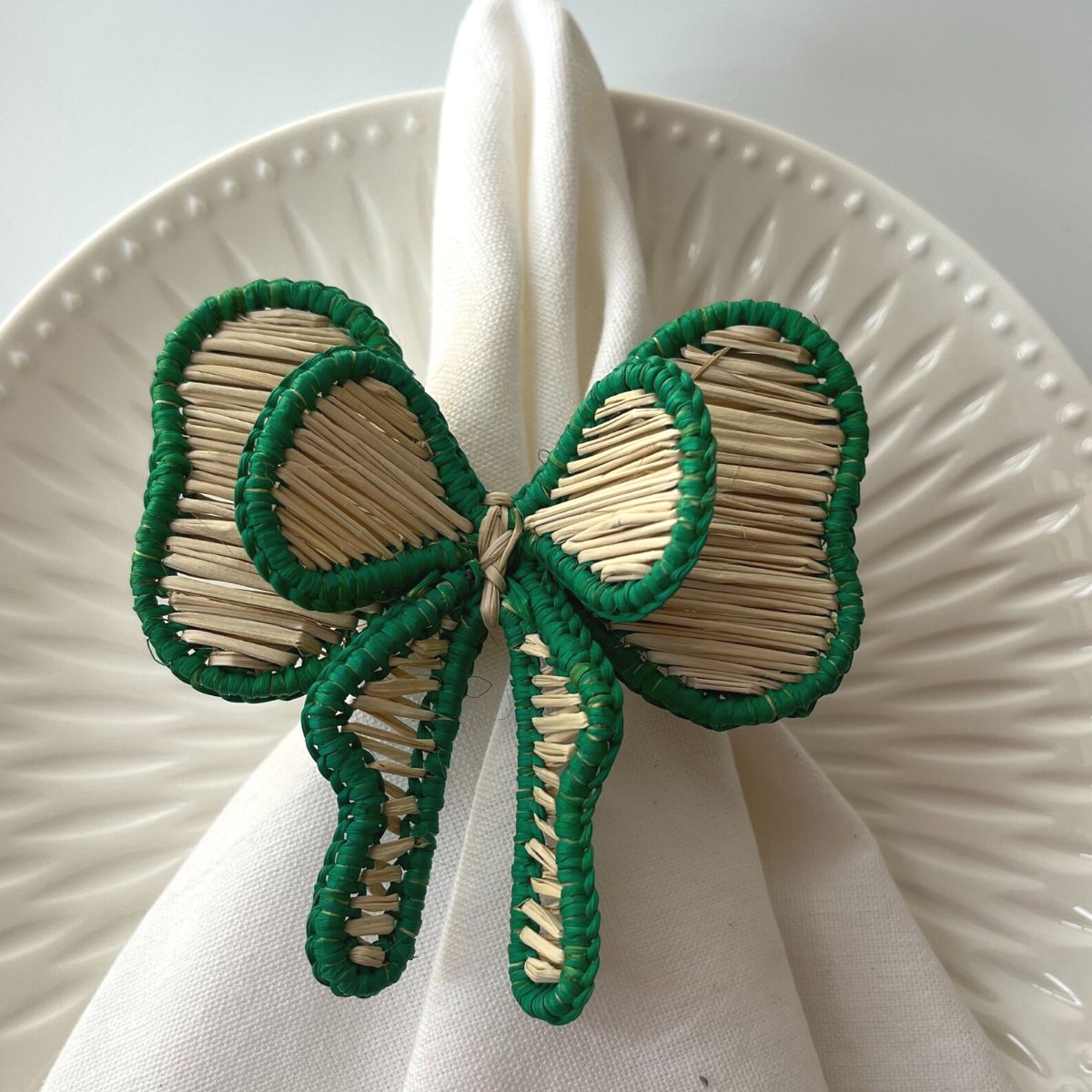 A green bow-shaped napkin ring made from natural iraca straw, displayed on a white plate.