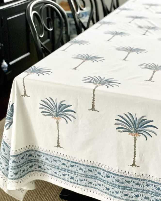 A palm tree blue hand block printed pattern on square tablecloth laid out on a table, with chairs around it and a window in the background.