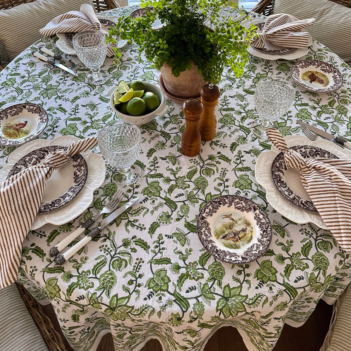 A set dining table with a green chintz hand block printed round tablecloth, featuring intricate floral pattern, surrounded by plates, cutlery, and striped napkins.