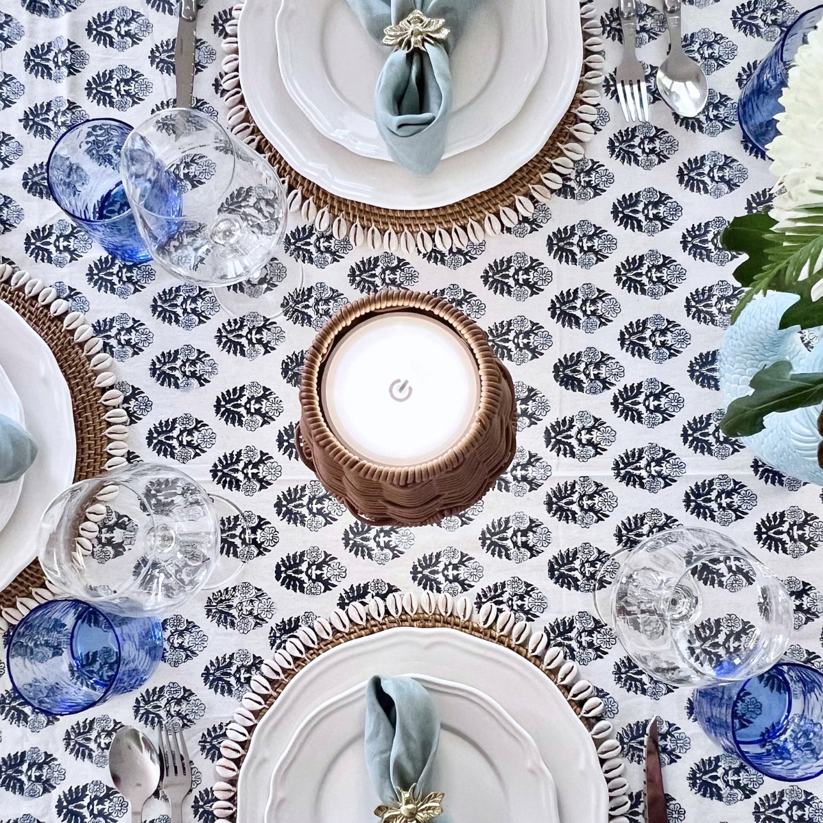 
Overhead view of a blue and white hand block printed tablecloth set for a special gathering with white plates, blue glasses and a woven candle lamp