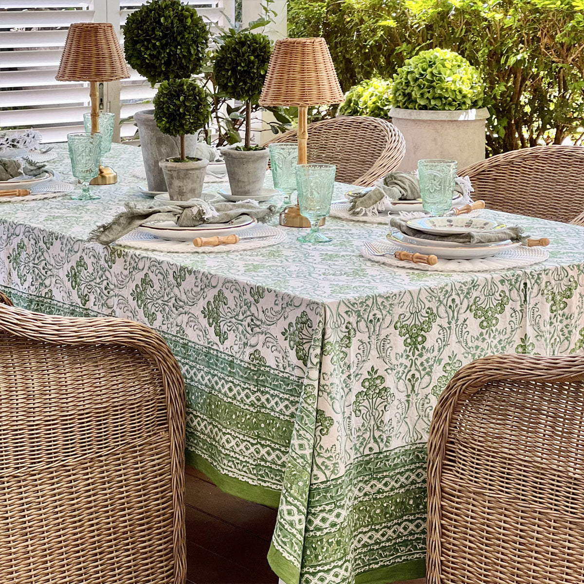 Rectangular green and white hand block printed tablecloth with an intricate border set for an outdoor lunch with topiary, rattan lamp and wicker chairs