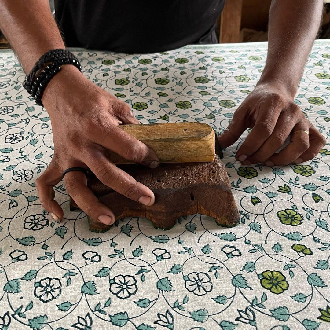 A craftsperson's hands pressing a carved wooden block onto premium cotton fabric, printing the Sea Breeze blue and green pattern.