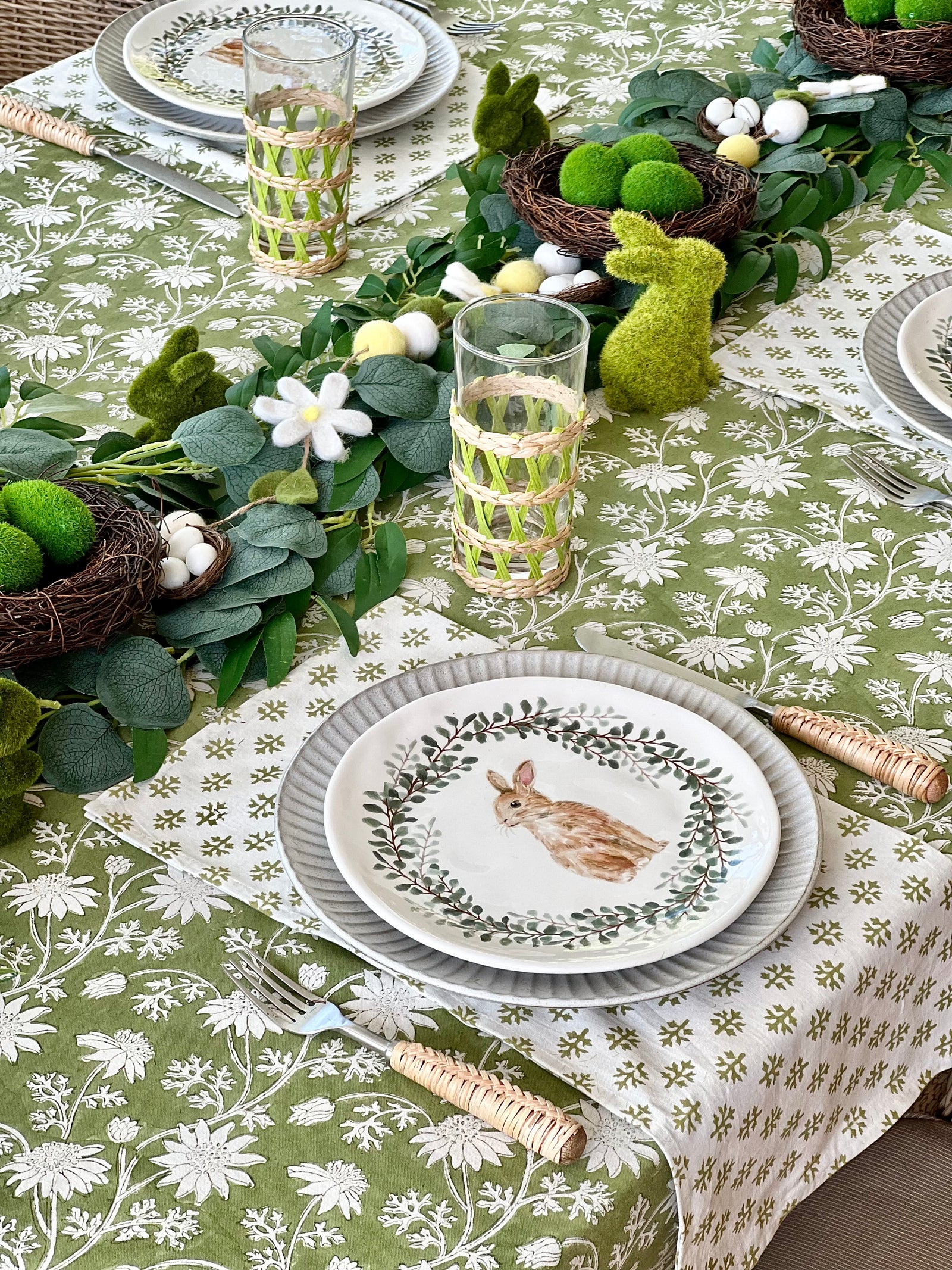 A set of hand block printed, green flannel foliage table cotton napkins placed on a table with floral tablecloth, next to a wicker placemat and a vase with lilies.