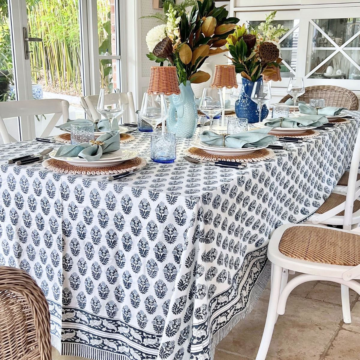 Rectangular blue floral hand block printed tablecloth in 180 x 270 cm on a dining table for eight to ten, with fresh flowers and woven chairs
