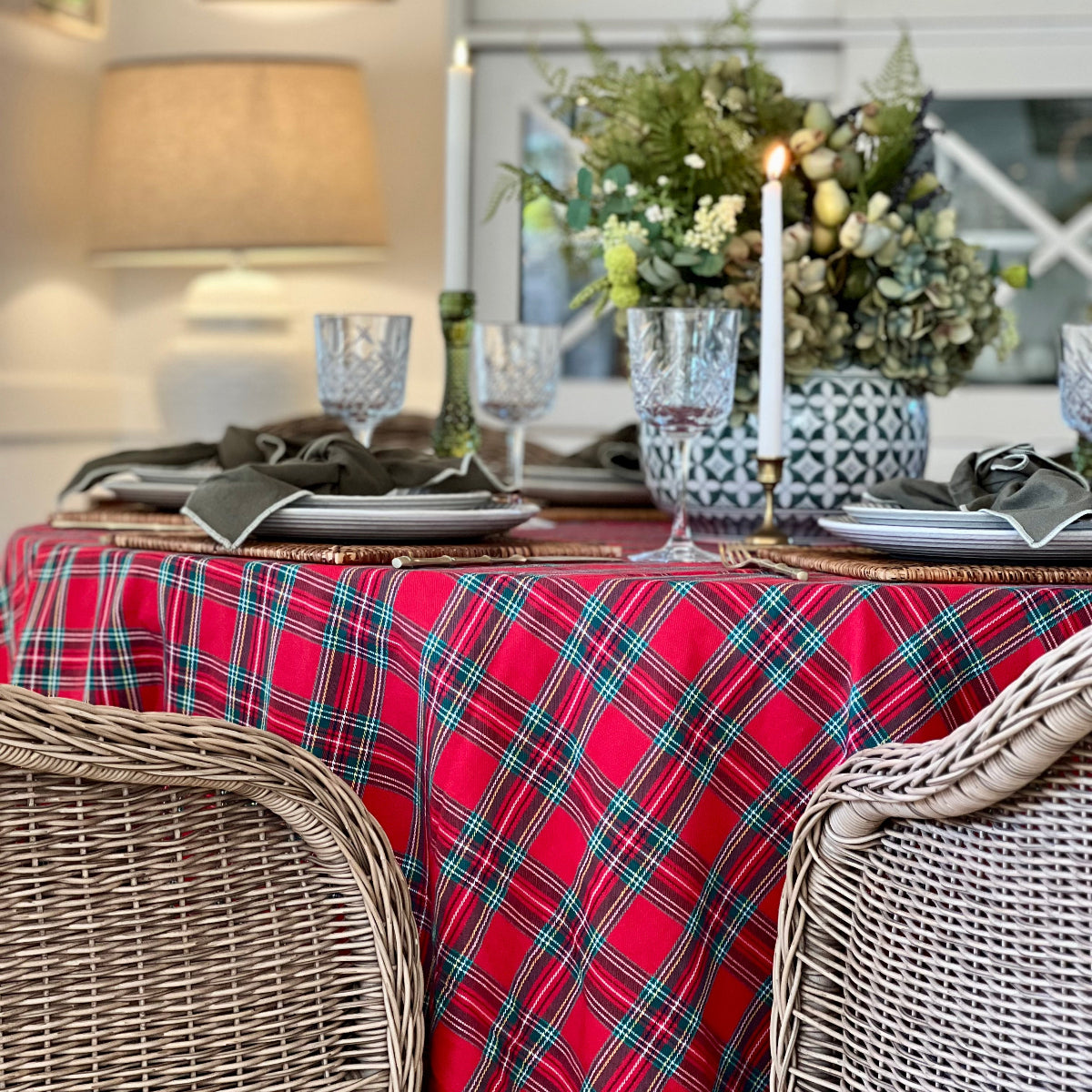 A red tartan plaid tablecloth with black,, white green and blue stripes, styled on a dining table with wicker chairs, glassware, candles, and a floral centerpiece in a cozy indoor setting.