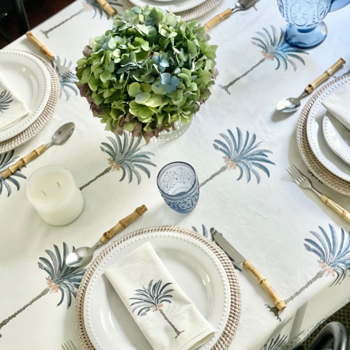 A hand block printed tablecloth with blue palm motifs on a white cotton base, displayed on a rectangular table with plates, cutlery, and a floral centerpiece.