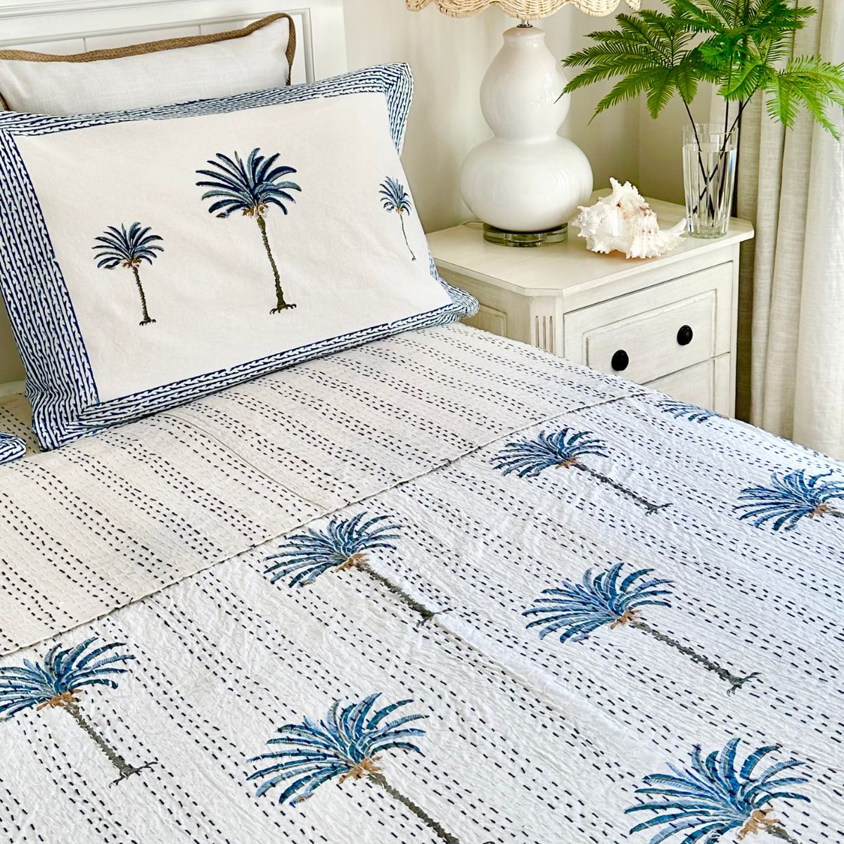 A hand block printed bedcover with a blue and white palm patterned Kantha quilt, with pillows matching the bedspread, in a room with light decor.