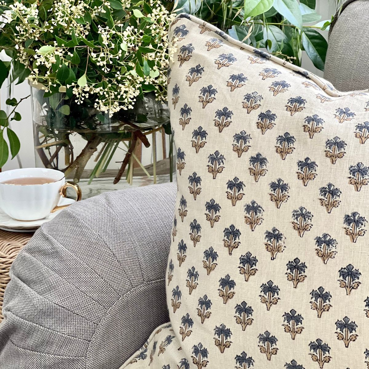 A 50 x 50 cm blue palm booti hand block printed pure linen cushion cover with pipe edge finish, styled upfront on a sofa with a coffee and floral vase.