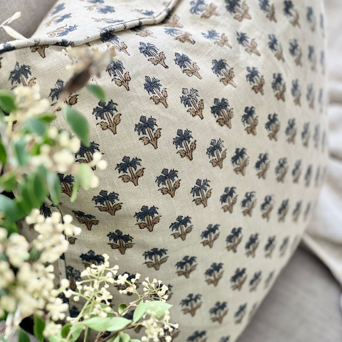 A 50 x 50 cm blue palm booti hand block printed pure linen cushion cover with pipe edge finish, styled upfront on a sofa with a coffee and floral vase.