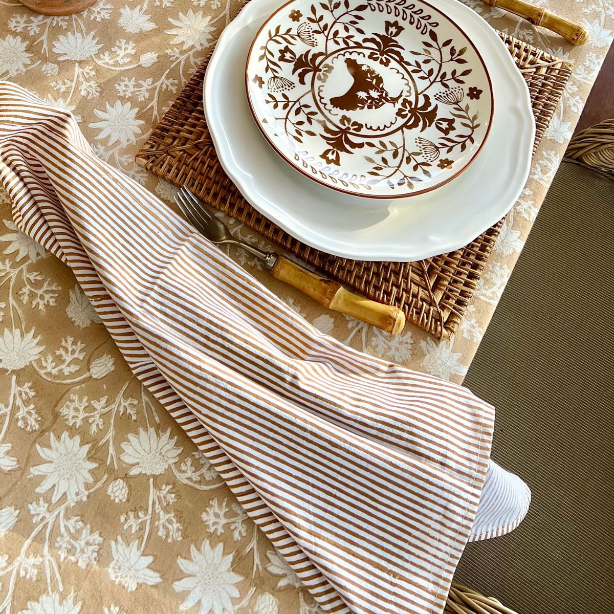 Single brown striped cotton napkin casually placed on a light brown tablecloth, creating a relaxed and inviting table setting for everyday dining.