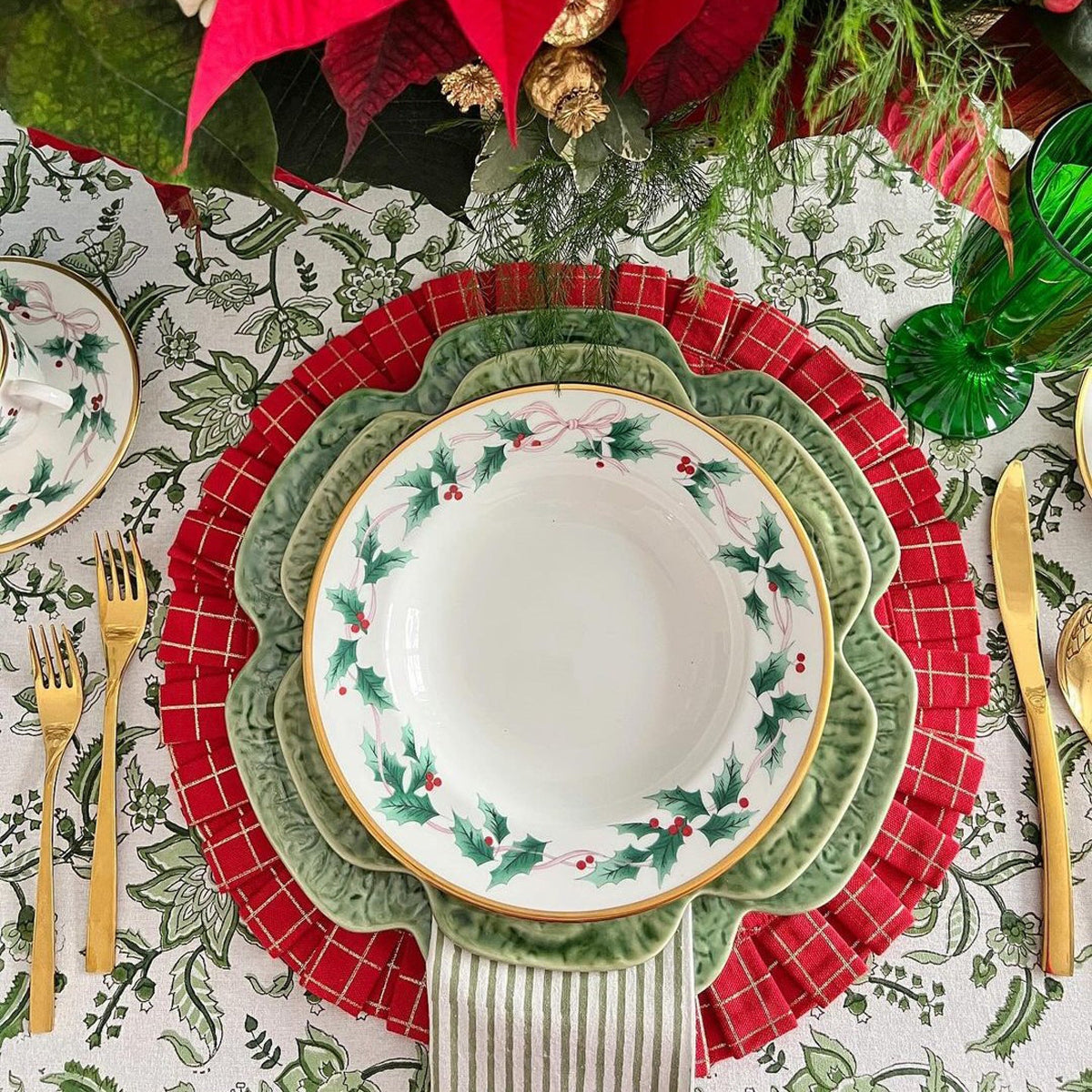 A Christmas-themed table setting featuring a large Chintz monochrome green hand block printed cotton table linen for a 10 to 12 seater table with decorative plates, red plaid placemats, and gold cutlery.