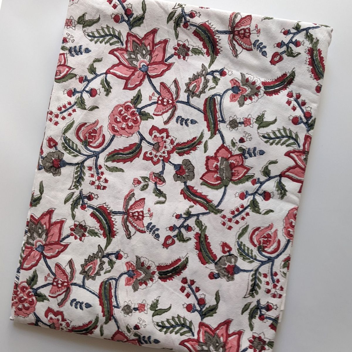 
A Chintz multicoloured hand block printed cotton square tablecloth folded flat, showing the vibrant pomegranate and flower pattern and complementary border on premium cotton fabric.