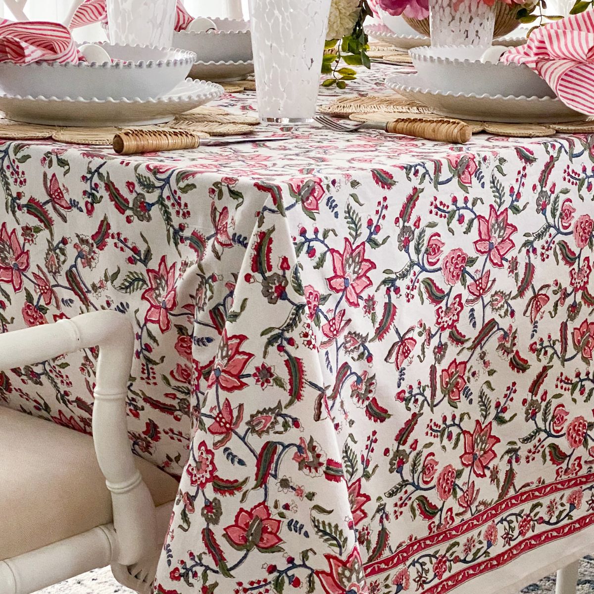 
A 150 x 220 cm Chintz multicoloured hand block printed cotton tablecloth on a 6 seater dining table, styled for a vibrant special occasion with white dishes, pink striped hand block printed napkins folded in a bow, and a floral centrepiece.