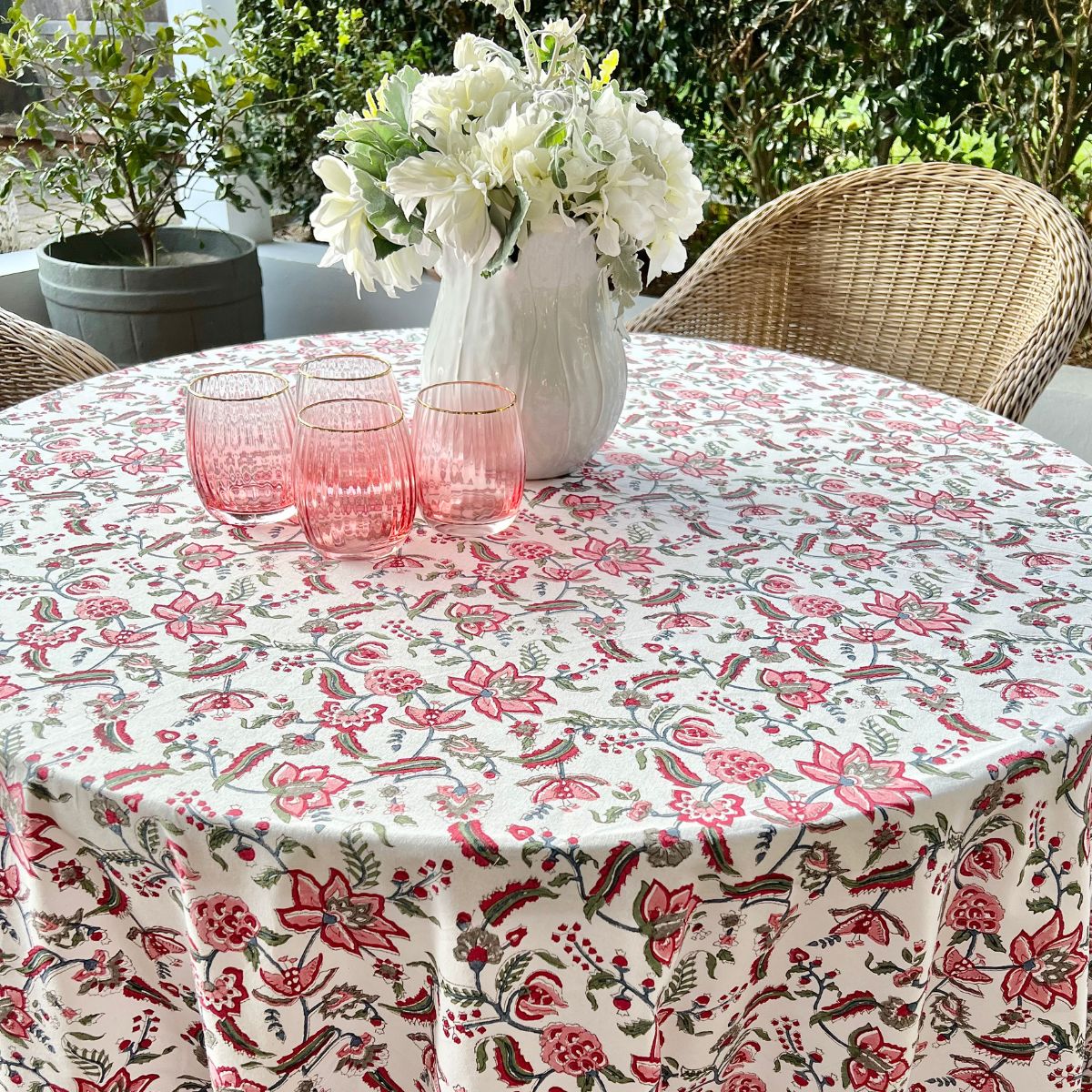 A multicolored round tablecloth with a pattern of hand block printed flowers, pomegranates and green leaves, displayed on a table set with plates and glasses.