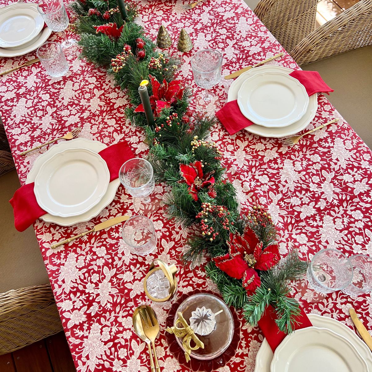 A red and white hand block printed floral patterned square tablecloth on a table with plates and cutlery set up for a meal.