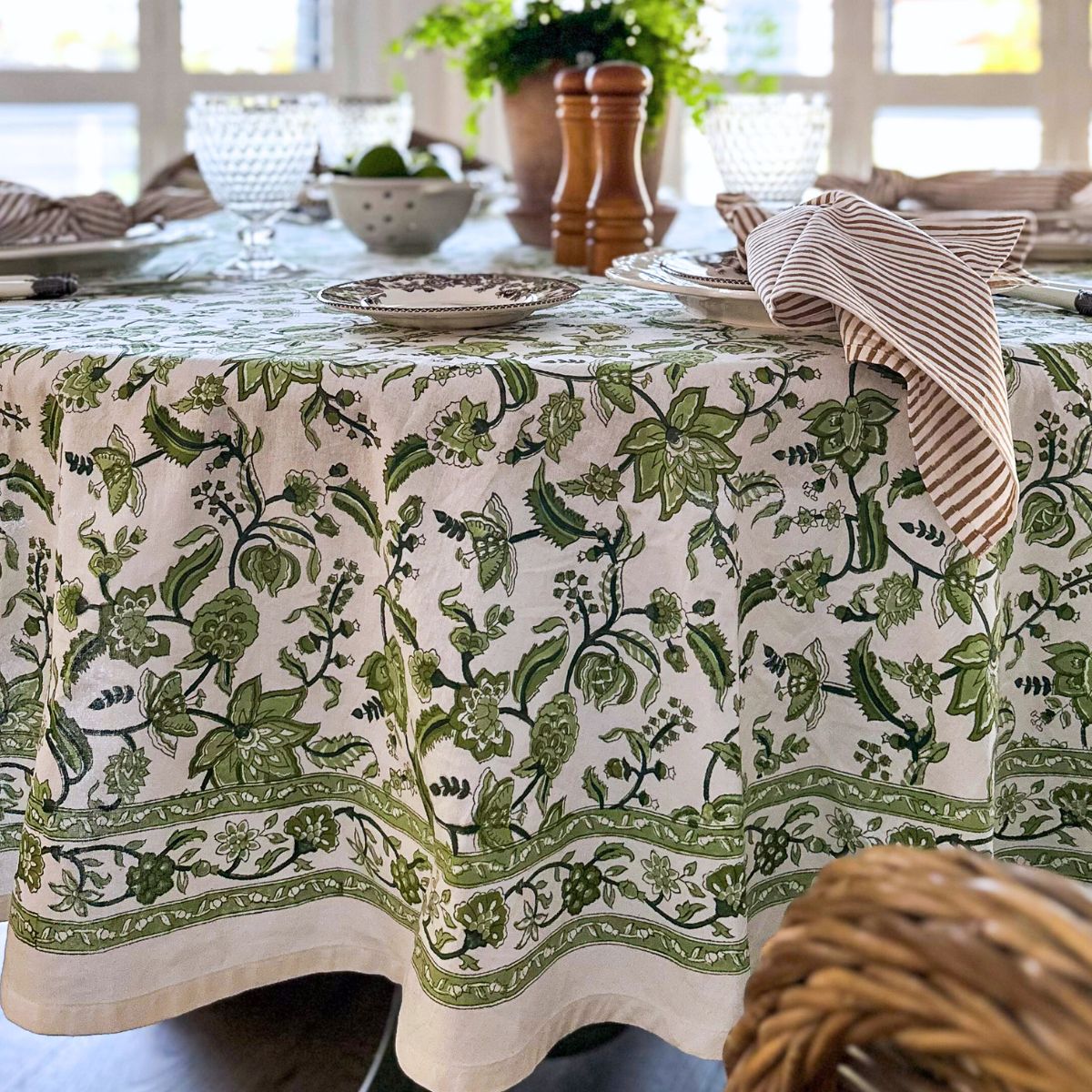 A set dining table with a green chintz hand block printed round tablecloth, featuring intricate floral pattern, surrounded by plates, cutlery, and striped napkins.