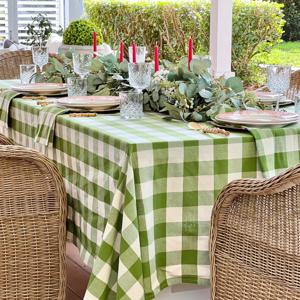 
A large green gingham check woven table linen on a 10 to 12 seater holiday dining table, featuring red decorative plates, red candles, eucalyptus and green foliage centrepiece, and green gingham napkins folded neatly beneath each plate, ideal for festive Australian Christmas entertaining.