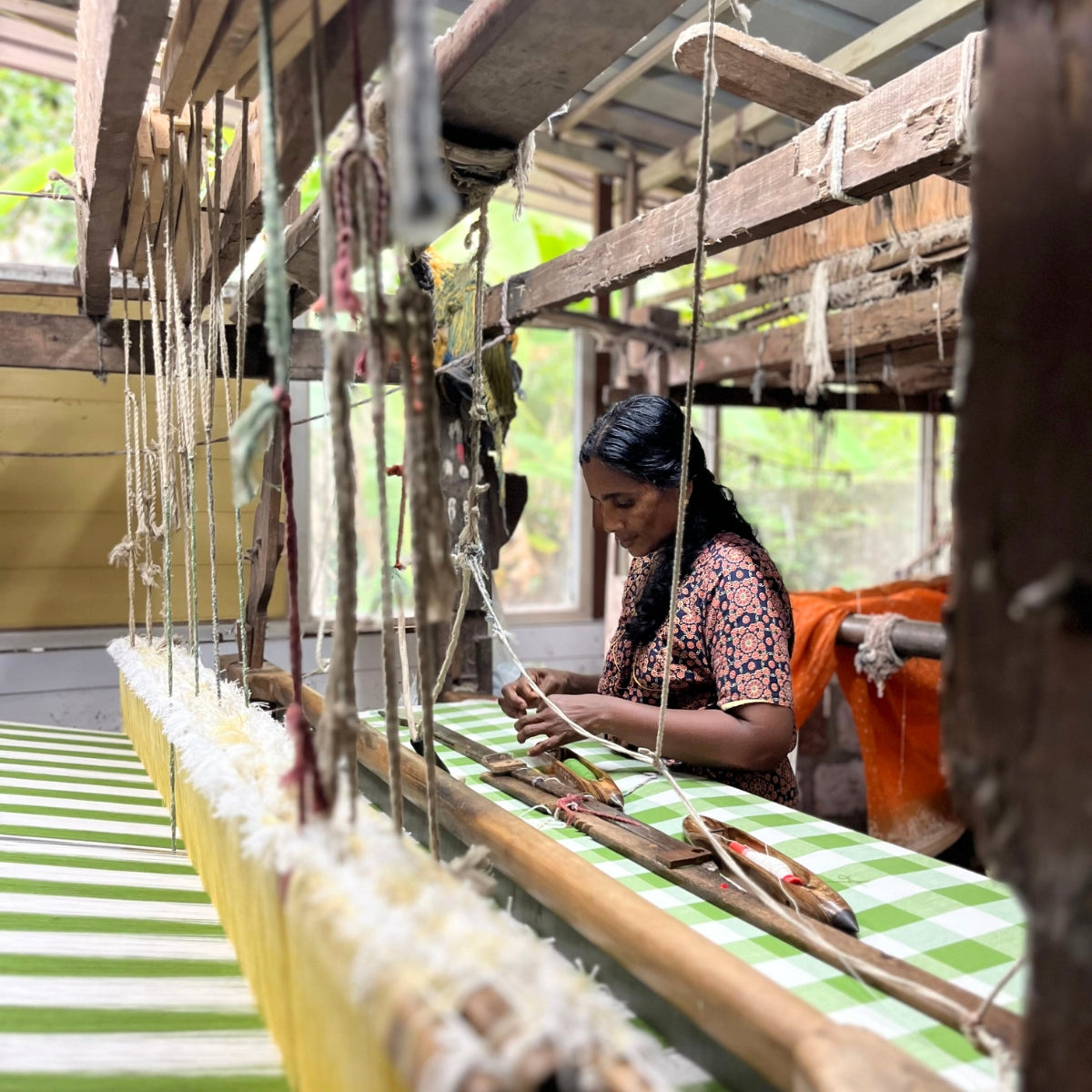 A skilled woman weaver at a traditional handloom in Kerala, India, weaving the green gingham check fabric carefully placed by hand in a small batch artisan process.