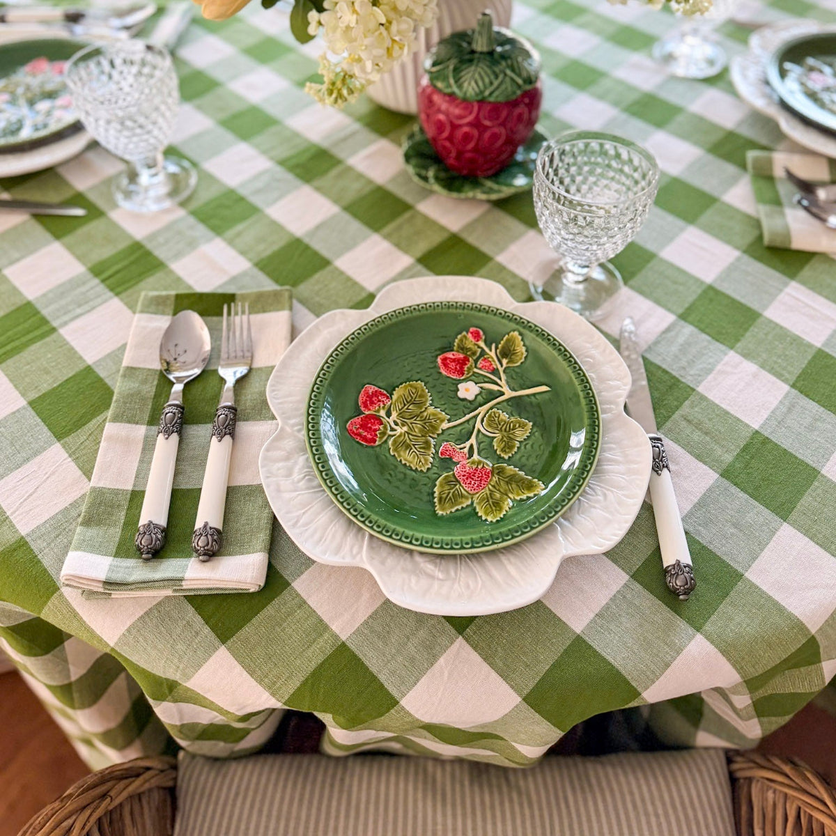 A large round green gingham check handwoven cotton tablecloth on a casual everyday dining table, featuring  decorative plates, folded green gingham napkins and a floral centrepiece for a fresh relaxed table setting.