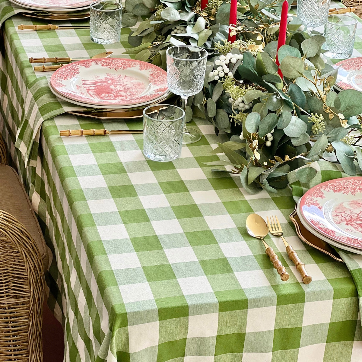 A green gingham handwoven tablecloth on a 6 to 8 seater Christmas table, styled with red decorative plates, bamboo cutlery, red taper candles, lush green foliage and eucalyptus along the centre, and green gingham napkins tucked beneath each place setting.