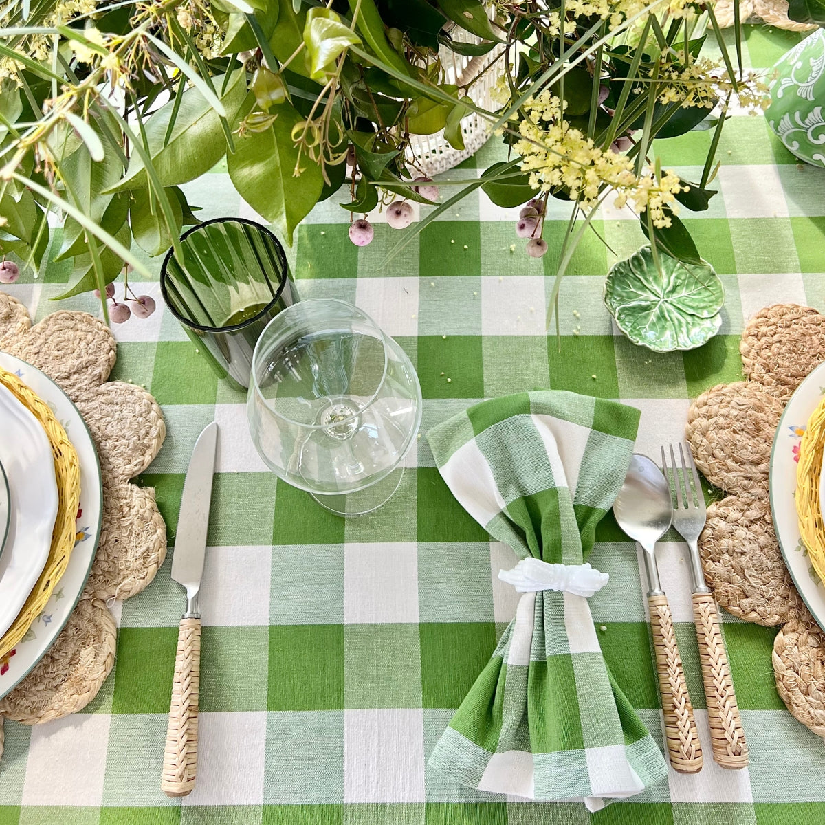A green gingham handwoven table linen on a square dining table set for everyday entertaining, featuring white decorative plates, bamboo cutlery, green gingham napkins, green glasses, and a fresh floral centrepiece.