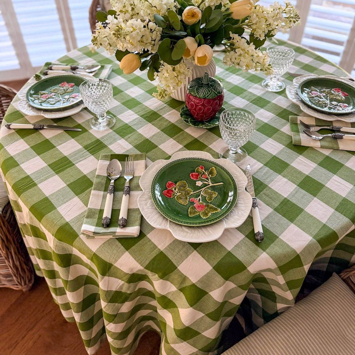 A 225 cm green gingham handwoven round table linen on an 8 seater circular table styled for a weekend brunch, with decorative plates, cutlery, green gingham napkins, glasses, and a fresh floral centrepiece.