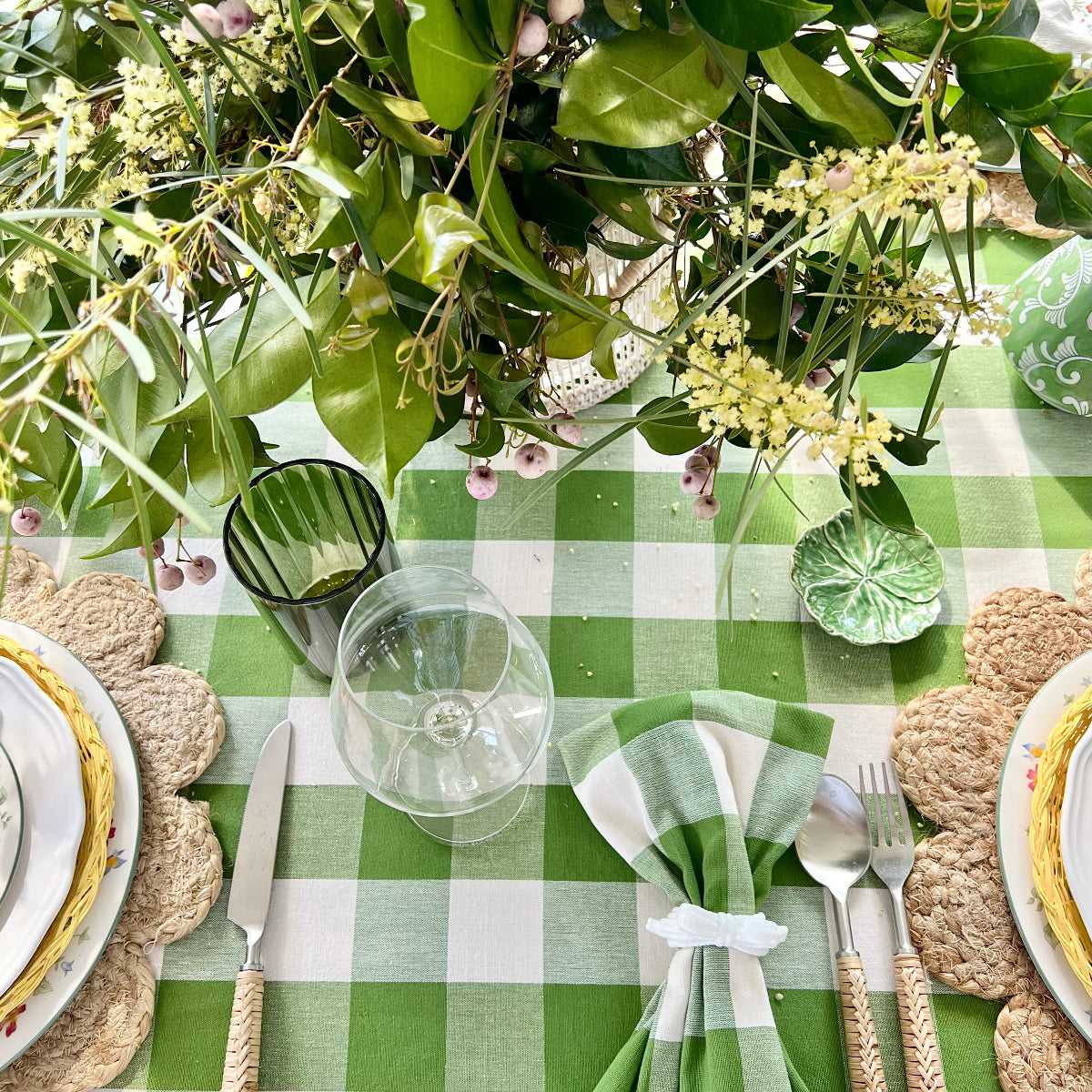 A green gingham check handwoven round tablecloth on an outdoor circular dining table set for alfresco entertaining, with white decorative plates, bamboo cutlery, green gingham napkins, green glasses, wine glasses, and a floral centrepiece, easy breezy Australian outdoor dining.