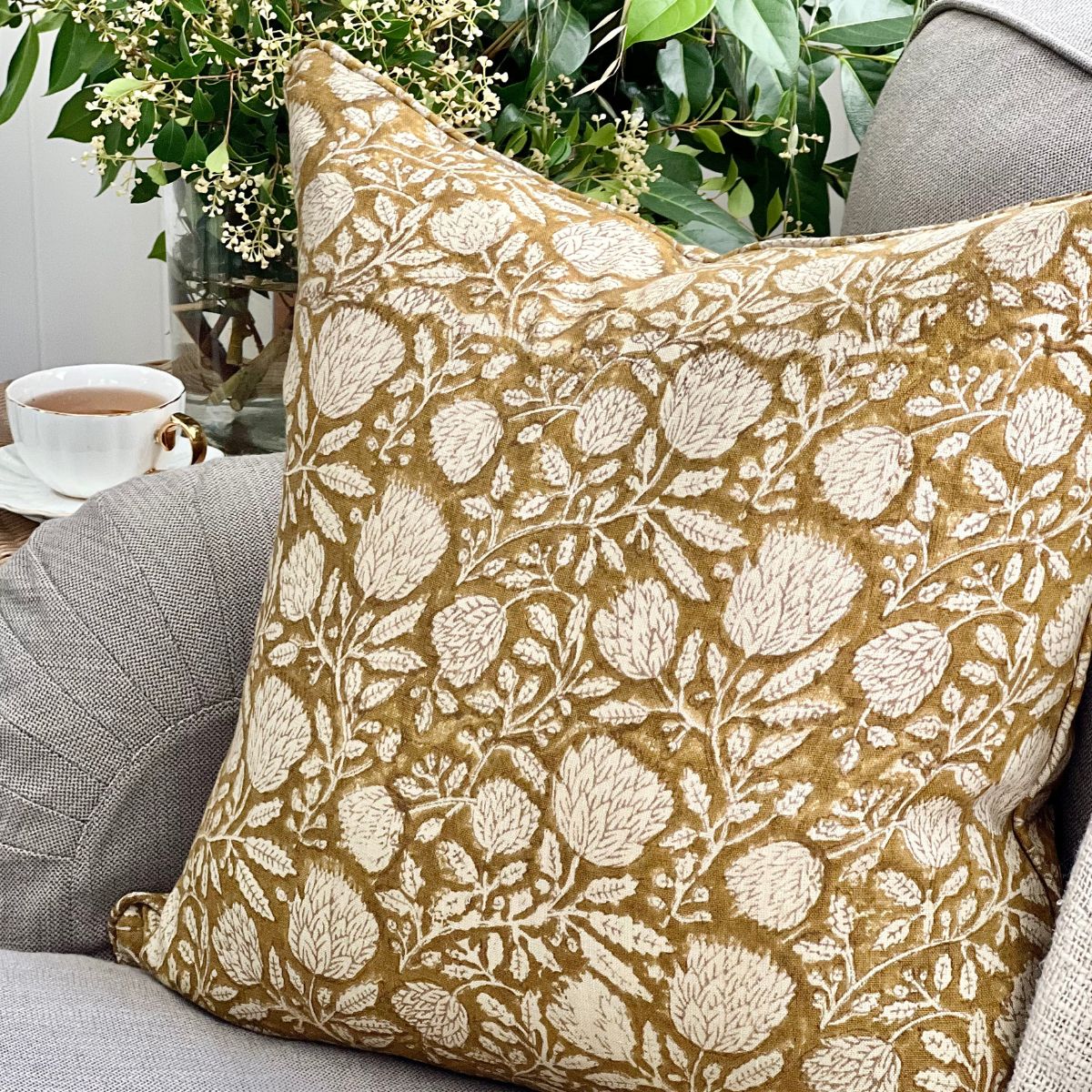 
A 50 x 50 cm Golden Flora mustard floral hand block printed pure linen cushion cover with pipe edge finish, styled upfront on a sofa with a coffee and floral vase.